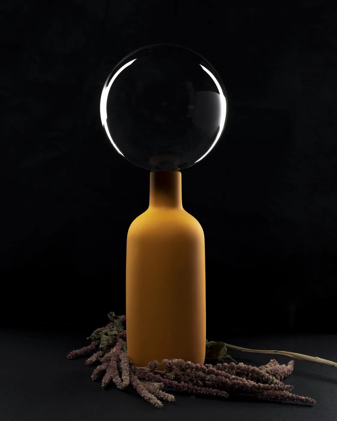 Ochre bottle topped with a glass sphere on a black ground. Object photography by STAS PRO