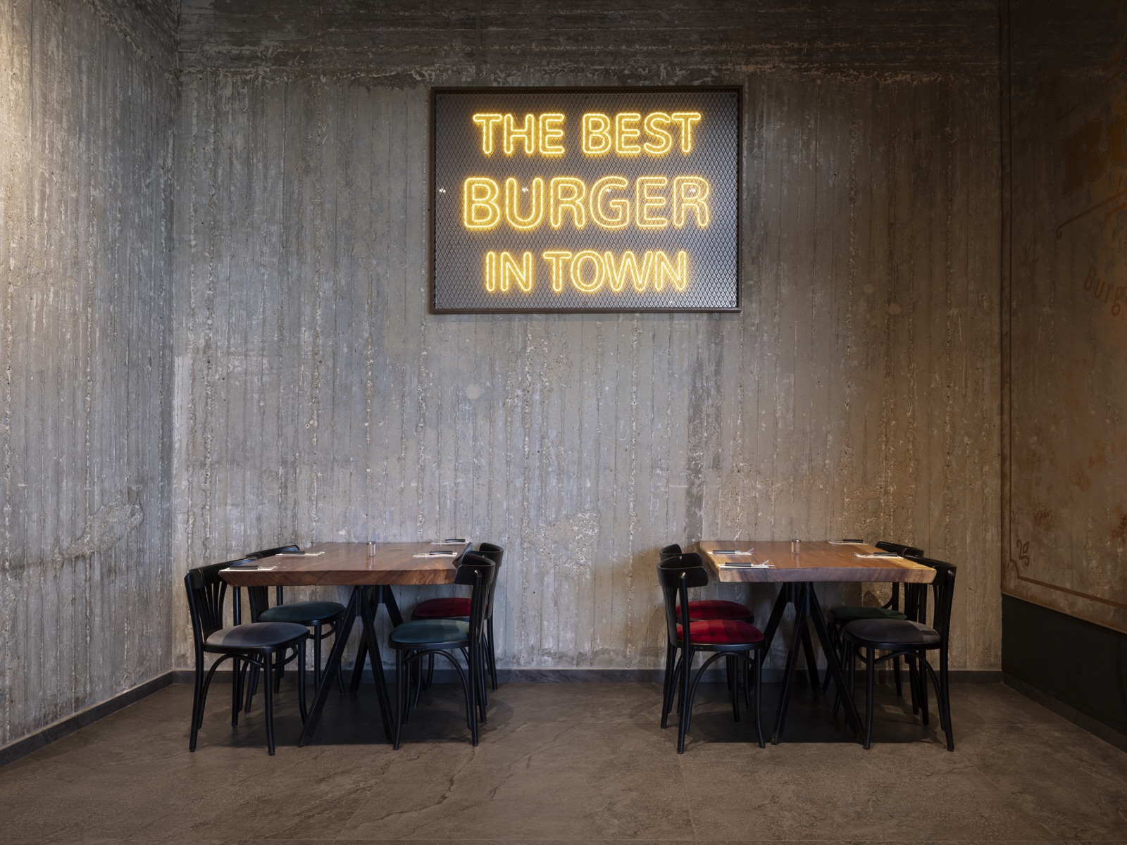 Neon sign reading The Best Burger in Town on a board-formed concrete wall above two wooden tables at Ruben restaurant, Ashkelon. Interior photography by STAS PRO